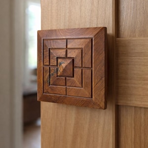 May include: A square, wooden door handle with a geometric design. The handle is a warm brown color and is mounted on a light-colored wooden door. The design features a series of squares and rectangles.
