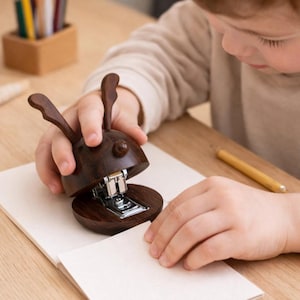May include: A dark brown wooden stapler in the shape of a rabbit. The stapler is open, revealing the metal stapling mechanism. The stapler is on top of a stack of white paper. A pencil and a wooden pencil holder are in the background.