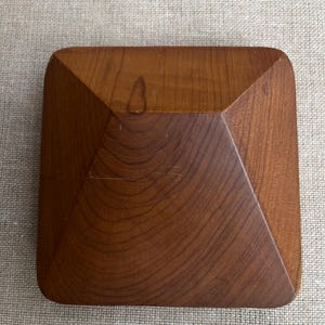 Puede incluir: Una caja cuadrada de madera con tapa piramidal. La caja es de madera de tono medio, con vetas visibles. Las esquinas son redondeadas y el diseño es simple y elegante. Ideal para guardar objetos.