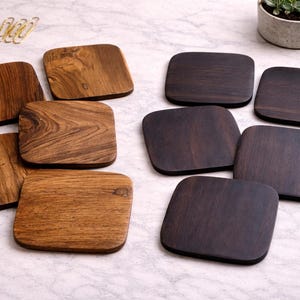 May include: A collection of wooden coasters in various shades of brown. The coasters have rounded corners and are arranged on a light-colored surface. Some coasters have a lighter wood grain, while others are a darker, richer brown.