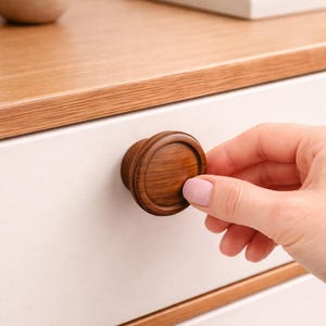 Puede incluir: Primer plano de un tirador de cajón de madera con un diseño redondo y en capas. El tirador es de color marrón cálido y está sujeto a un cajón blanco. El cajón forma parte de un mueble de madera.