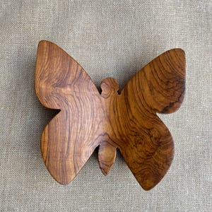 May include: A wooden butterfly sculpture with a natural wood grain pattern. The butterfly is a warm brown color and has detailed wing shapes. The sculpture is placed on a neutral-colored, textured surface.