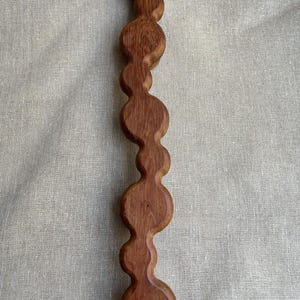 May include: A handcrafted wooden decorative piece with a series of connected, rounded shapes. The wood has a rich, reddish-brown hue and visible grain. The design is vertical and symmetrical, suggesting a unique home decor item.