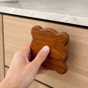 May include: A wooden cabinet drawer pull with a unique, organic shape. The pull is a warm, medium brown color and appears to be made of solid wood. The drawer is made of light-colored wood. The pull is being held by a hand.