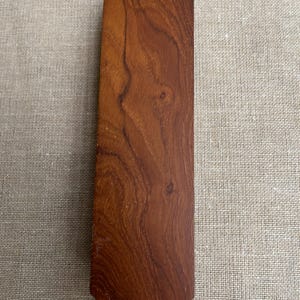 May include: A solid, wooden tie with a pointed bottom and a flat top. The tie is a rich, reddish-brown color with visible wood grain patterns. The tie is displayed on a neutral, textured background.