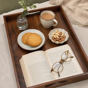 May include: A rectangular dark brown wooden serving tray with cutout handles. The tray holds a clear glass vase with a green sprig, a ceramic mug with a light brown beverage, a small plate with two round cookies, a scalloped bowl of nuts, and an open book with a pair of brown-rimmed glasses resting on the pages.
