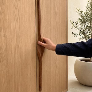 May include: A long, vertical, dark brown wooden cabinet handle with a twisted, organic shape mounted on a light oak wood cabinet door. The handle features a smooth, contoured center section that tapers toward the top and bottom ends.