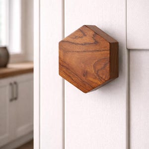 Puede incluir: Un pomo hexagonal de madera, con acabado marrón cálido, montado en una puerta blanca. La forma geométrica y la veta natural de la madera crean una estética moderna y minimalista. Adecuado para armarios de cocina o baño.