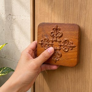 May include: A square, wooden cabinet knob with a fleur-de-lis design. The knob is a warm brown color and is held by a hand. The background includes a wooden door and a glimpse of a plant.