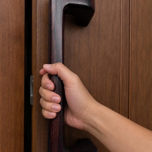 May include: A dark brown wooden door handle with a curved design. The handle is being held by a person's hand. The door and frame are made of wood with a similar color.