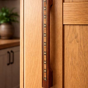 May include: A wooden cabinet pull with a dark brown finish and inlaid decorative accents. The pull features a vertical design with a central strip of alternating diamond and rectangular patterns in lighter wood and brass. The pull is mounted on a light-colored wooden cabinet door.