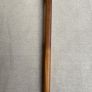 Könnte beinhalten: Ein massives Nudelholz aus Holz mit einem glatten, abgerundeten Griff. Das Nudelholz hat eine warme, mittelbraune Farbe und scheint aus Hartholz zu bestehen. Der Griff ist leicht gebogen. Für den Küchengebrauch.