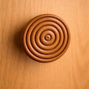 May include: A round, wooden cabinet knob with a spiral design. The knob is a warm brown color and has a series of concentric rings. The knob is mounted on a light brown wooden surface.