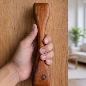 May include: A vertical wooden door handle with a smooth, contoured shape and a natural brown wood grain finish. The handle features a distinct dark knot detail near the bottom and is mounted onto a matching light brown wooden door surface.