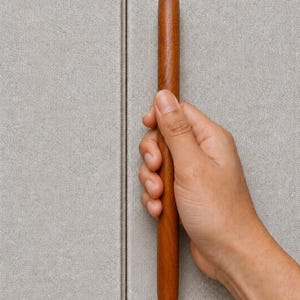 May include: A hand holding a cylindrical, wooden cabinet pull. The pull is a warm brown color and has a smooth, rounded shape. The background is a light gray, textured surface.