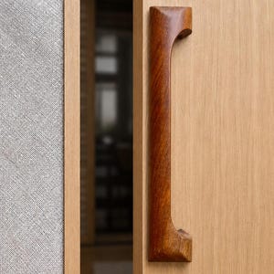 May include: A vertical, dark brown wooden door handle with a smooth, curved profile mounted on a light-colored wooden door panel. The handle features a solid, polished finish with visible wood grain, positioned next to a textured gray panel.