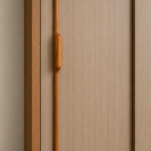 May include: A long, vertical, light brown wooden cabinet handle with a smooth, rounded central grip section, mounted on a light oak-colored cabinet door with a visible wood grain texture and a vertical sliding panel design.