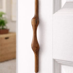 May include: A handcrafted wooden door pull with a unique, organic shape. The pull is made of a warm-toned wood, mounted on a white door. The design features a series of rounded sections connected by slender areas, creating an elegant, sculptural form.