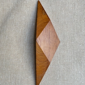 Könnte beinhalten: Ein geometrisches Holzobjekt mit einer rautenförmigen Mitte und dreieckigen Enden. Das Holz hat einen warmen, rötlich-braunen Farbton und sichtbare Maserung. Das Objekt befindet sich vor einem neutralen, strukturierten Hintergrund.