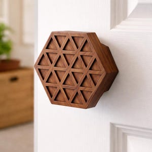 Puede incluir: Un pomo de puerta hexagonal de madera con un patrón triangular geométrico. El pomo es de color marrón cálido y está montado en una puerta blanca. El diseño es moderno y decorativo.