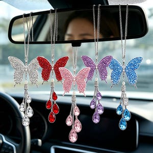 Op de afbeelding: Vijf vlinder auto-ornamenten die aan een achteruitkijkspiegel hangen. Elk ornament heeft een vlinderontwerp met strass in zilver, rood, roze, paars en blauw. Elk ornament heeft een hangend druppelontwerp.