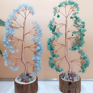May include: Two decorative gemstone trees. One tree features light blue stones, the other green. Both have copper wire branches and trunks, set in wooden bases filled with colorful stones. The background is a neutral beige.