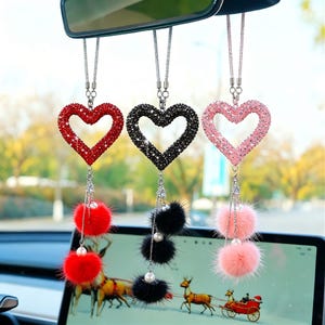 Peut inclure: Trois breloques de voiture en forme de cœur, rouges, noires et roses, ornées de strass étincelants. Chaque breloque est suspendue à une chaîne argentée et comporte un pompon duveteux et une fausse perle. Les breloques sont suspendues au rétroviseur d'une voiture.