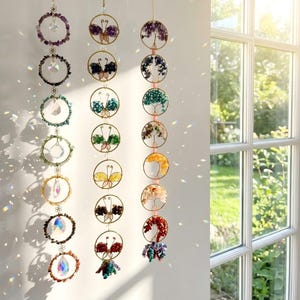 May include: A collection of hanging suncatchers with circular frames and various gemstone and crystal designs. The suncatchers feature butterfly and tree of life motifs, with a range of colors including purple, green, and red. They are suspended by gold chains.
