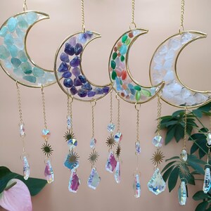 Op de afbeelding: Vier maanvormige zonneweerders met goudkleurige frames, elk gevuld met kleurrijke edelstenen. Elke maan hangt aan een ketting en heeft bungelende kristallen en sterrencharms. De edelstenen zijn blauw, paars, groen en wit.