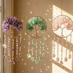 Op de afbeelding: Drie hangende decoratieve zonnevangers met een levensboomontwerp. Elk heeft een cirkelvormig goudkleurig frame met een boom gemaakt van amethist, groene en roze stenen. Strengen kralen en kristallen hangen eronder.