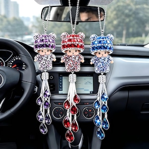 Op de afbeelding: Drie auto-hangornamenten met een klein poppenontwerp. Elke pop heeft een hoed en outfit versierd met sprankelende kristallen in paars, rood en blauw. Elk ornament heeft een zilveren ketting en een kwast van druppelvormige kristallen.