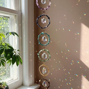 May include: A hanging chakra crystal suncatcher with seven colorful gemstone rings. Each ring features a different color of gemstones, including purple, blue, green, yellow, brown, and orange. Clear crystal prisms hang from each ring, reflecting light.