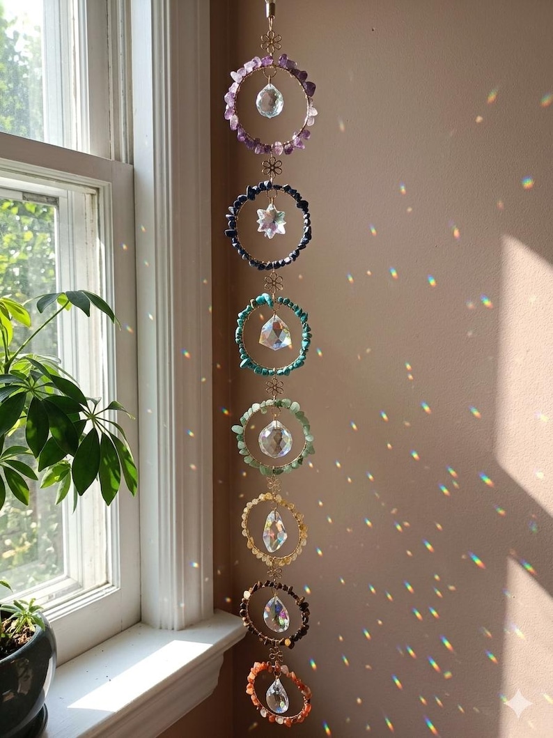 May include: A hanging chakra crystal suncatcher with seven colorful gemstone rings. Each ring features a different color of gemstones, including purple, blue, green, yellow, brown, and orange. Clear crystal prisms hang from each ring, reflecting light.
