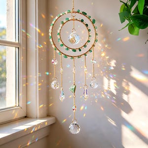 May include: A decorative suncatcher with two gold-colored rings, adorned with green beads and clear crystals. Clear crystal pendants hang from the rings, creating a prism effect. The suncatcher is suspended in front of a window.