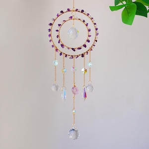 May include: A gold-colored dream catcher with two concentric rings adorned with purple stones. Clear crystal beads and iridescent accents hang from the bottom, creating a decorative and ethereal aesthetic. The dream catcher is suspended from a thin gold chain.
