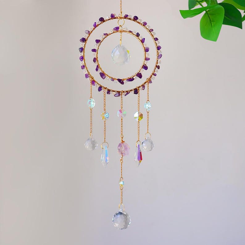 May include: A gold-colored dream catcher with two concentric rings adorned with purple stones. Clear crystal beads and iridescent accents hang from the bottom, creating a decorative and ethereal aesthetic. The dream catcher is suspended from a thin gold chain.