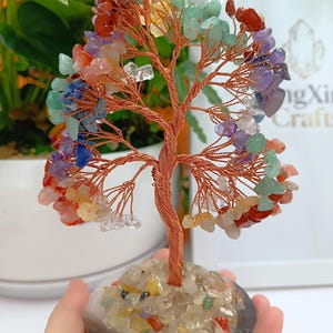 May include: A decorative tree sculpture with a copper wire frame and branches, decorated with colourful gemstones. The base is made of small, polished stones. The tree is held in a hand, with a white background.