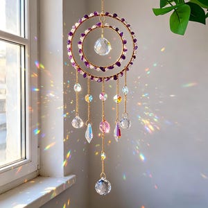 May include: A decorative suncatcher with two gold-colored rings adorned with purple and clear stones. Clear crystal pendants and beads hang from the rings, reflecting light. The suncatcher is suspended by a gold chain, creating a beautiful display.