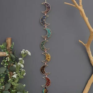 May include: A decorative wall hanging featuring six crescent moon shapes in a vertical line. Each gold-colored moon is filled with colorful stones and connected by pink beads. The bottom moon has a cluster of stones.