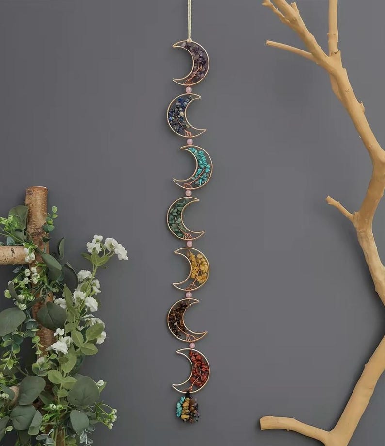 May include: A decorative wall hanging featuring six crescent moon shapes in a vertical line. Each gold-colored moon is filled with colorful stones and connected by pink beads. The bottom moon has a cluster of stones.