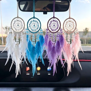 Op de afbeelding: Vier dromenvangers hangen aan de achteruitkijkspiegel van een auto. Elk heeft een cirkelvormig frame met kralen en veren in wit, blauw, paars en roze. De dromenvangers hangen aan een koord in bijpassende kleur.