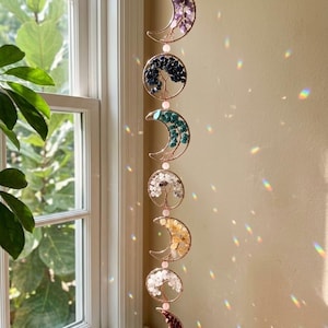 May include: A hanging moon phase suncatcher with six crescent moon and circle designs. Each piece features a different colored gemstone arrangement, including purple, blue, and yellow. The suncatcher is suspended by a thin, off-white ribbon.