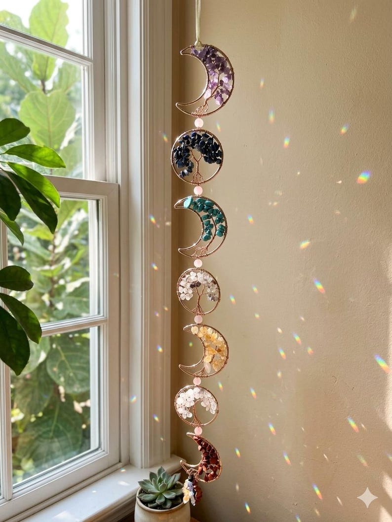May include: A hanging moon phase suncatcher with six crescent moon and circle designs. Each piece features a different colored gemstone arrangement, including purple, blue, and yellow. The suncatcher is suspended by a thin, off-white ribbon.