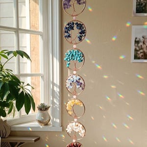 May include: A hanging, decorative mobile featuring seven circular frames with wire tree designs. Each circle is filled with colorful gemstones, including purple, blue, turquoise, white, yellow, and red. The mobile hangs in front of a window.