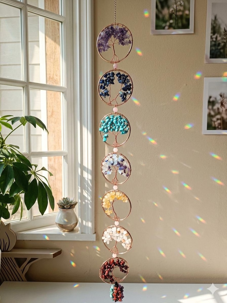 May include: A hanging, decorative mobile featuring seven circular frames with wire tree designs. Each circle is filled with colorful gemstones, including purple, blue, turquoise, white, yellow, and red. The mobile hangs in front of a window.