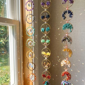 May include: A collection of hanging crystal suncatchers and gemstone ornaments. The suncatchers feature colorful crystals and gemstones in various shapes and sizes, including butterflies and tree of life designs. The ornaments are suspended by gold-colored chains.