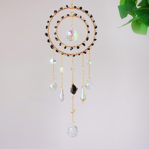 May include: A decorative suncatcher with a gold-colored metal frame in a double-circle design. The circles are adorned with black beads. Clear and iridescent crystal pendants hang from the frame, with a large crystal ball in the center.