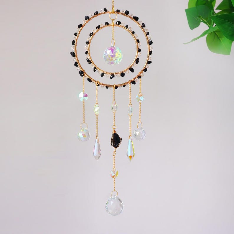 May include: A decorative suncatcher with a gold-colored metal frame in a double-circle design. The circles are adorned with black beads. Clear and iridescent crystal pendants hang from the frame, with a large crystal ball in the center.