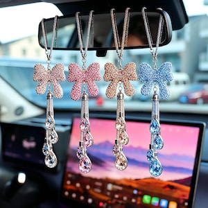 Op de afbeelding: Vier auto-achteruitkijkspiegelbedels met strikvormige toppen en bungelende kristallen druppels. De strikken zijn in tinten roze, goud en blauw, elk bedekt met sprankelende strass-steentjes. De bedels hangen aan zilveren kettingen.