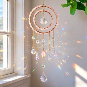 May include: A decorative suncatcher with a double-ring design. The rings are adorned with pink beads and gold-colored metal. Clear crystal pendants and faceted crystals hang from the rings, creating a prism effect. The suncatcher is hanging near a window.